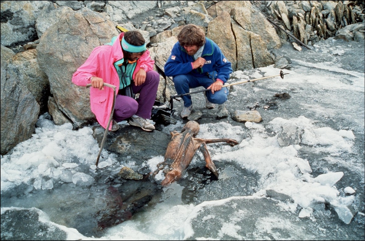 Two people in outdoor gear sit on the edge of where ice and rock meet, staring at a decomposed body.